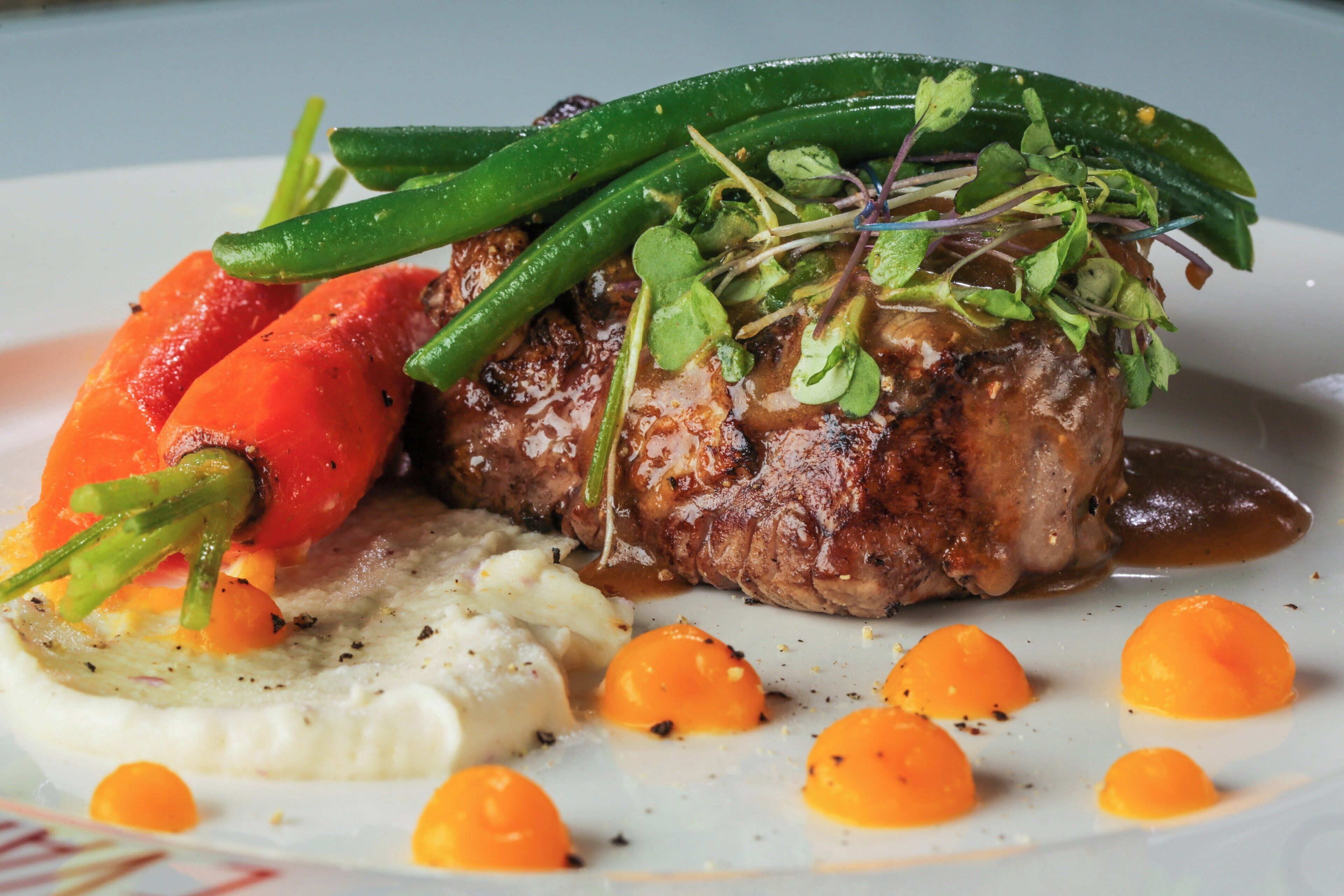 A plated dish with a steak, green beans, carrots, and a creamy side.