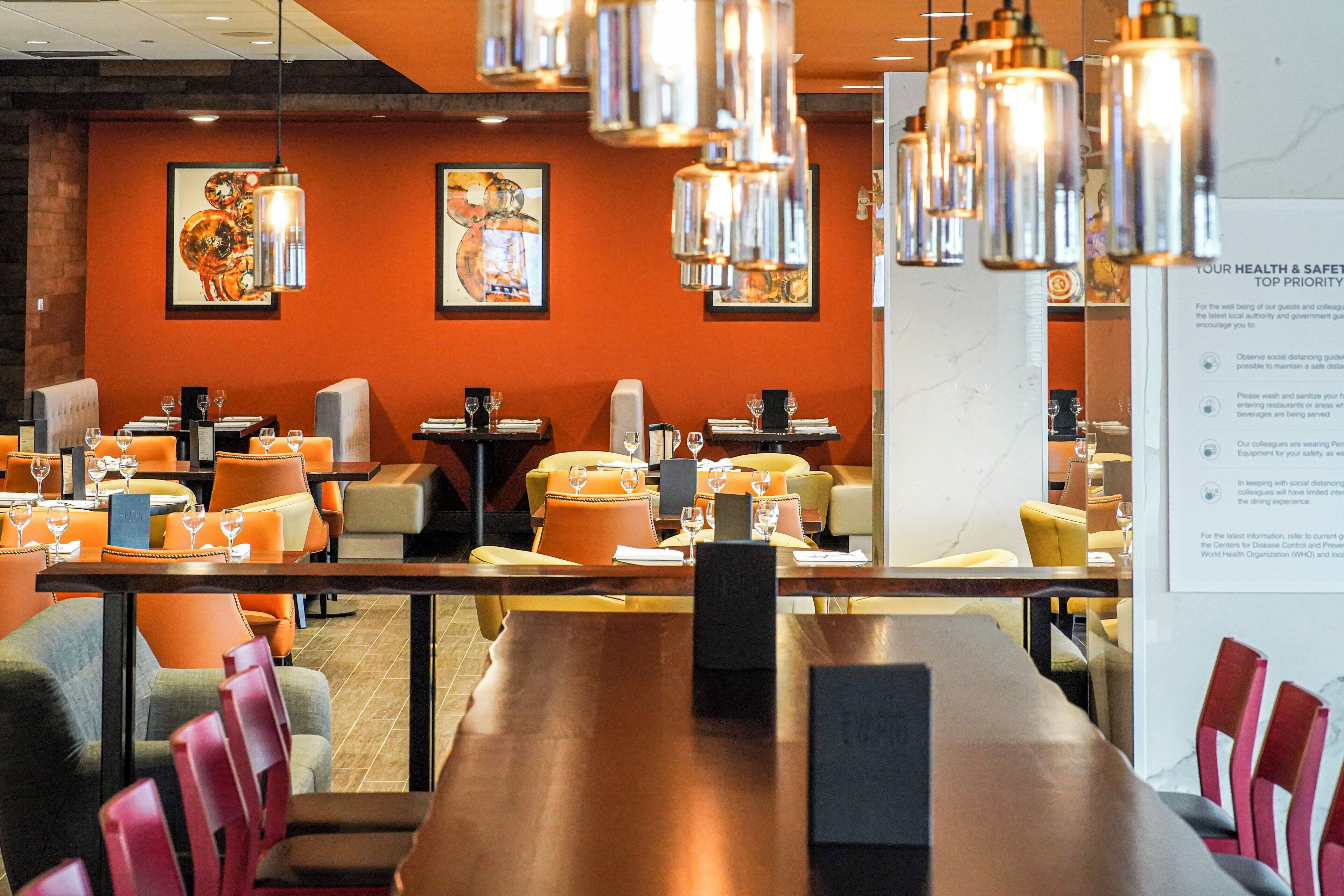 Un restaurante con varias mesas y sillas, algunas con cojines naranjas. Hay lámparas colgantes y cuadros en las paredes.