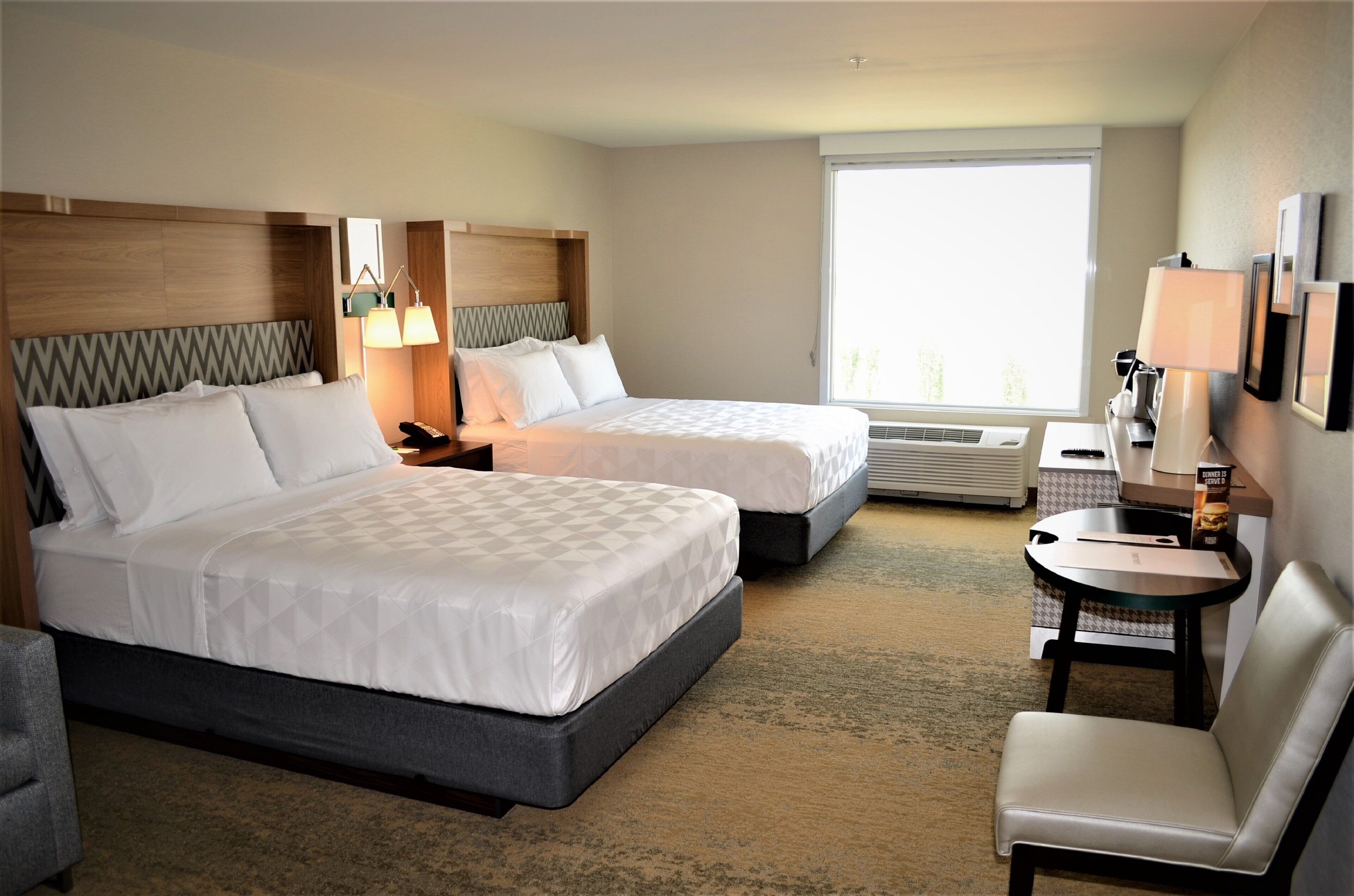 A hotel room with two beds, a desk, a chair, a window, and a wall-mounted lamp.