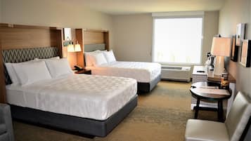 A hotel room with two beds, a desk, a chair, a window, and a wall-mounted lamp.