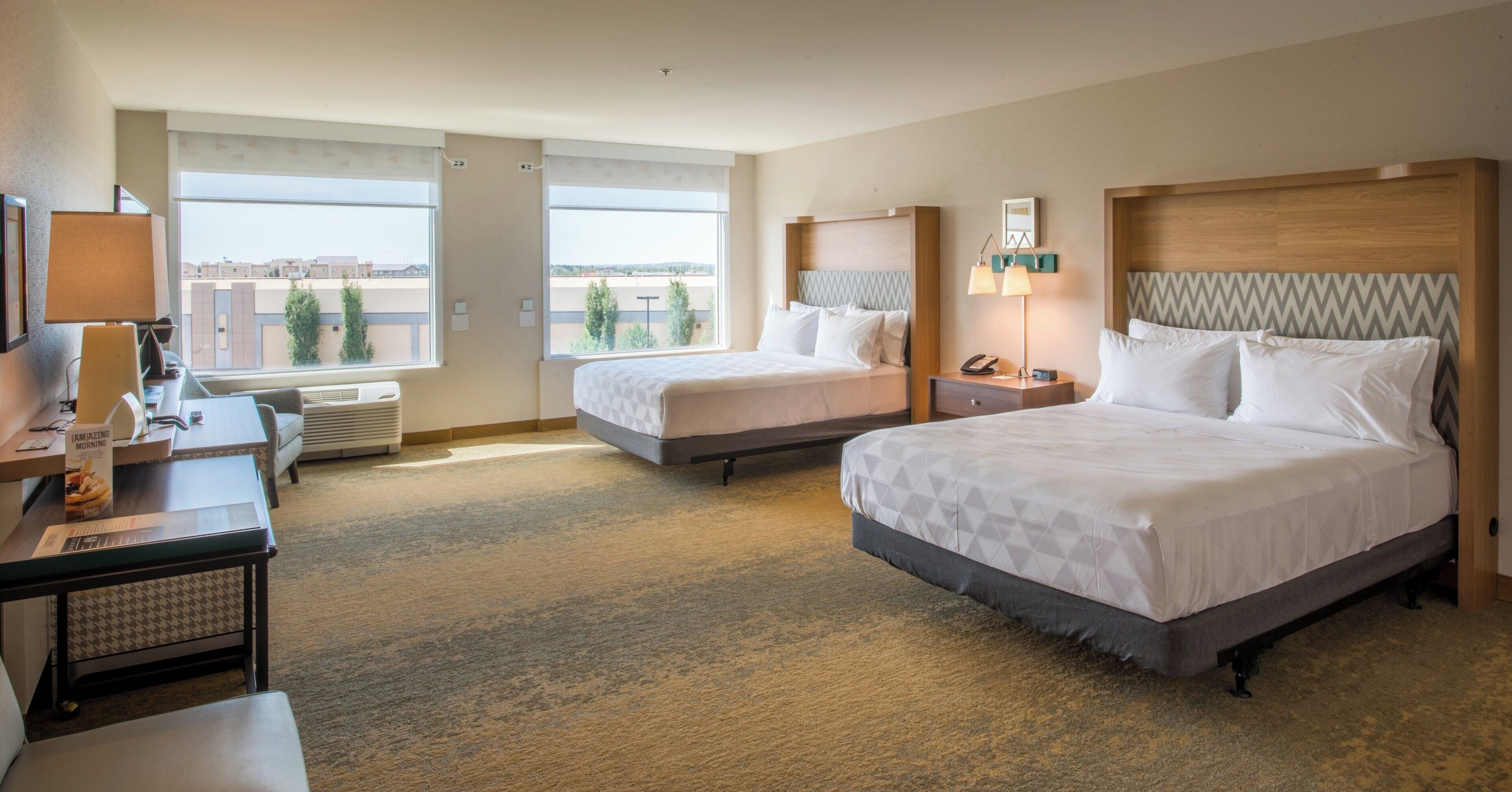 A hotel room with two beds, a desk, a chair, a lamp, and a view of the city.