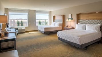 A hotel room with two beds, a desk, a chair, a lamp, and a view of the city.