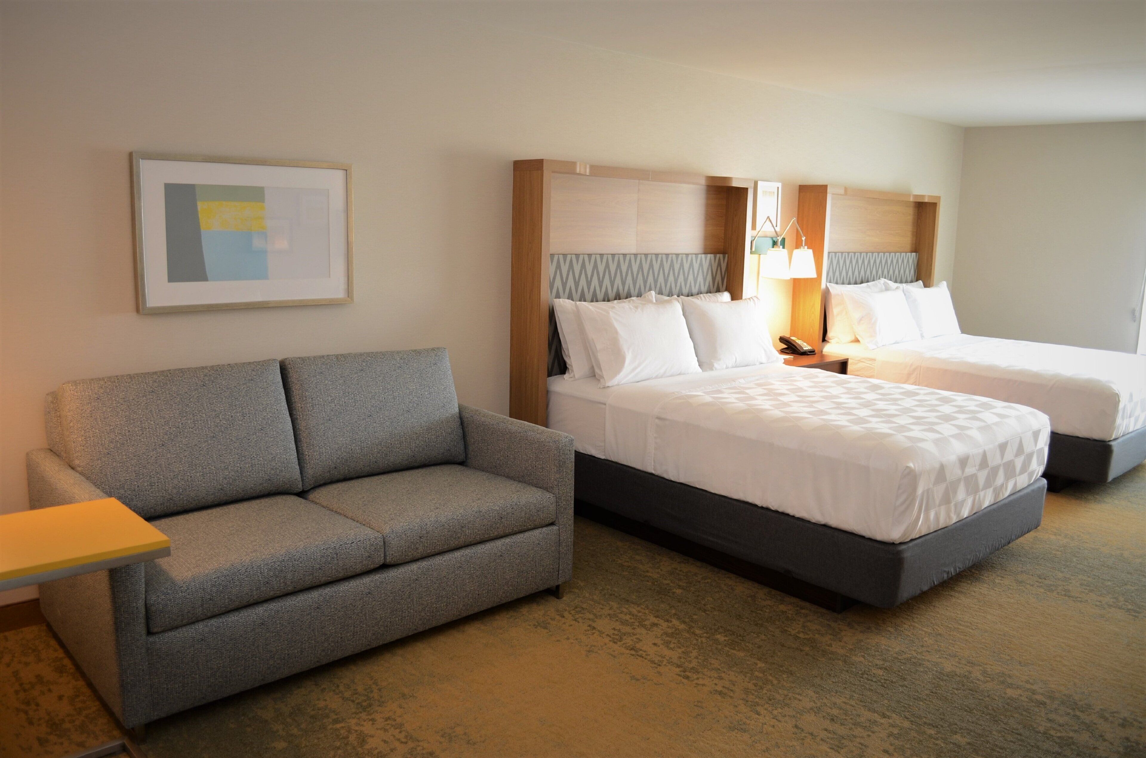 A hotel room with a grey sofa, two beds with white bedding, a framed picture on the wall, and a bedside lamp.
