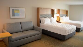 A hotel room with a grey sofa, two beds with white bedding, a framed picture on the wall, and a bedside lamp.