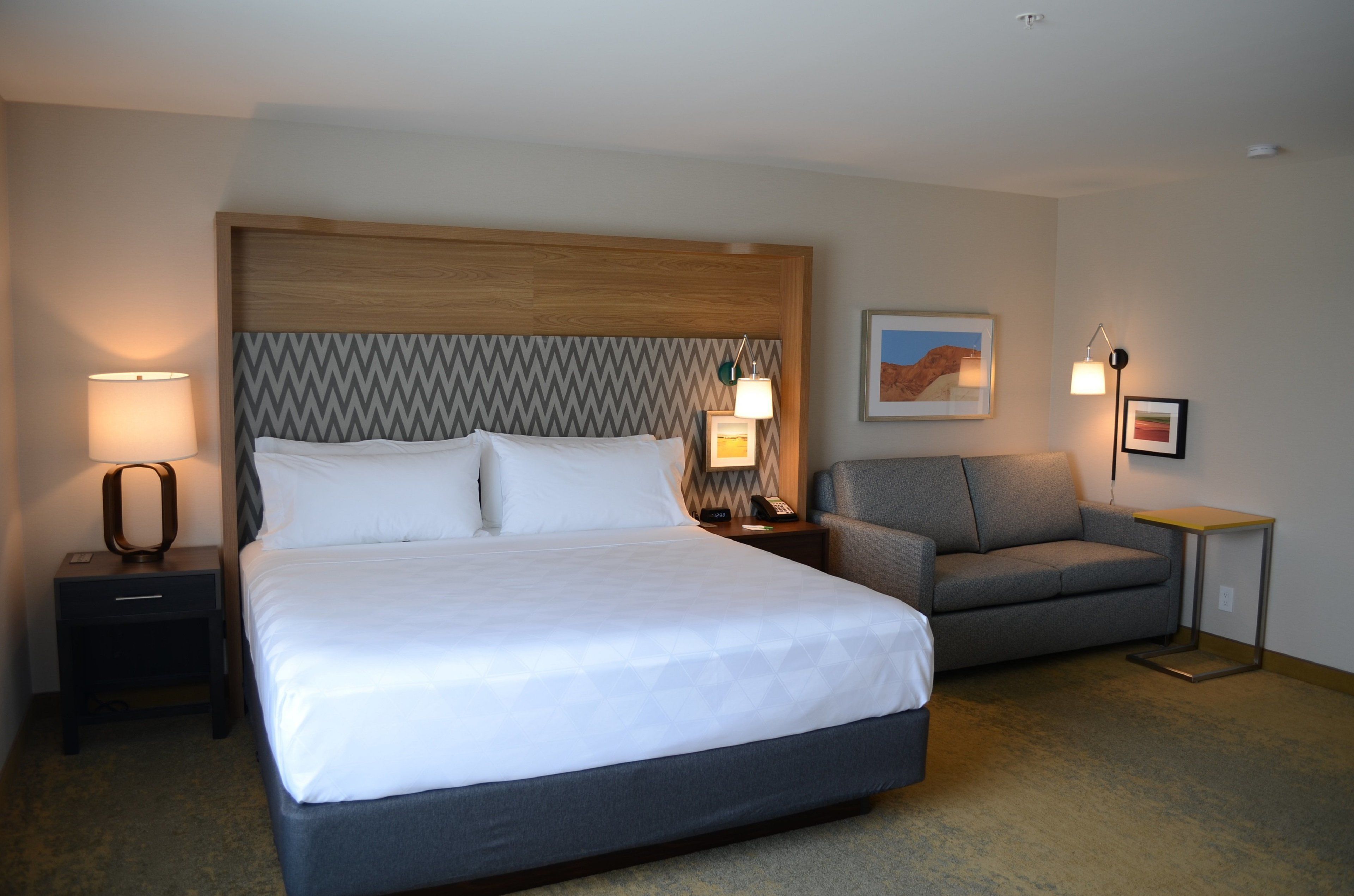 A hotel room with a large bed, a grey sofa, a wooden headboard, and two bedside lamps.