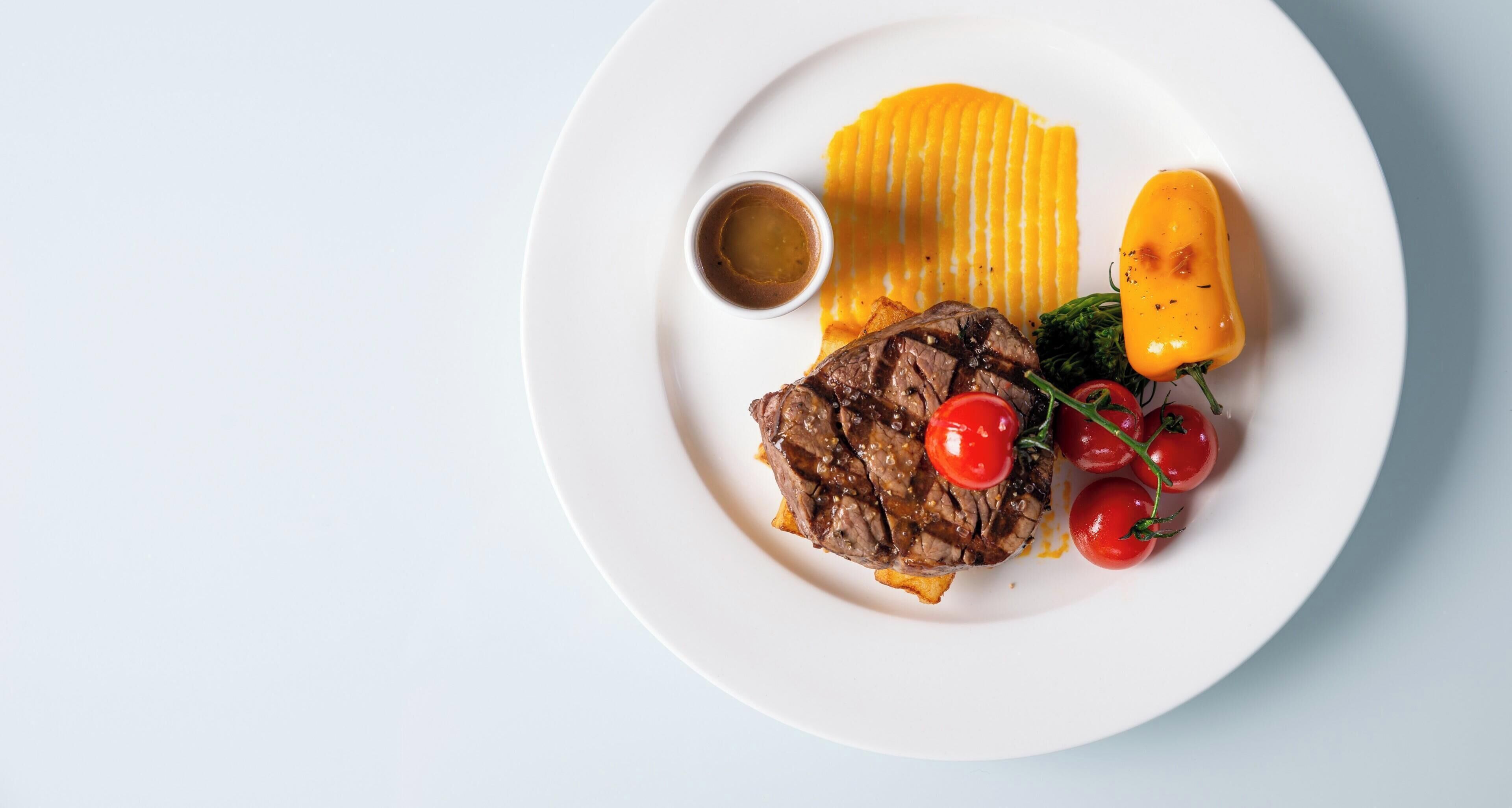 A plate with grilled steak, grilled yellow bell pepper, cherry tomatoes, and a small bowl of sauce.