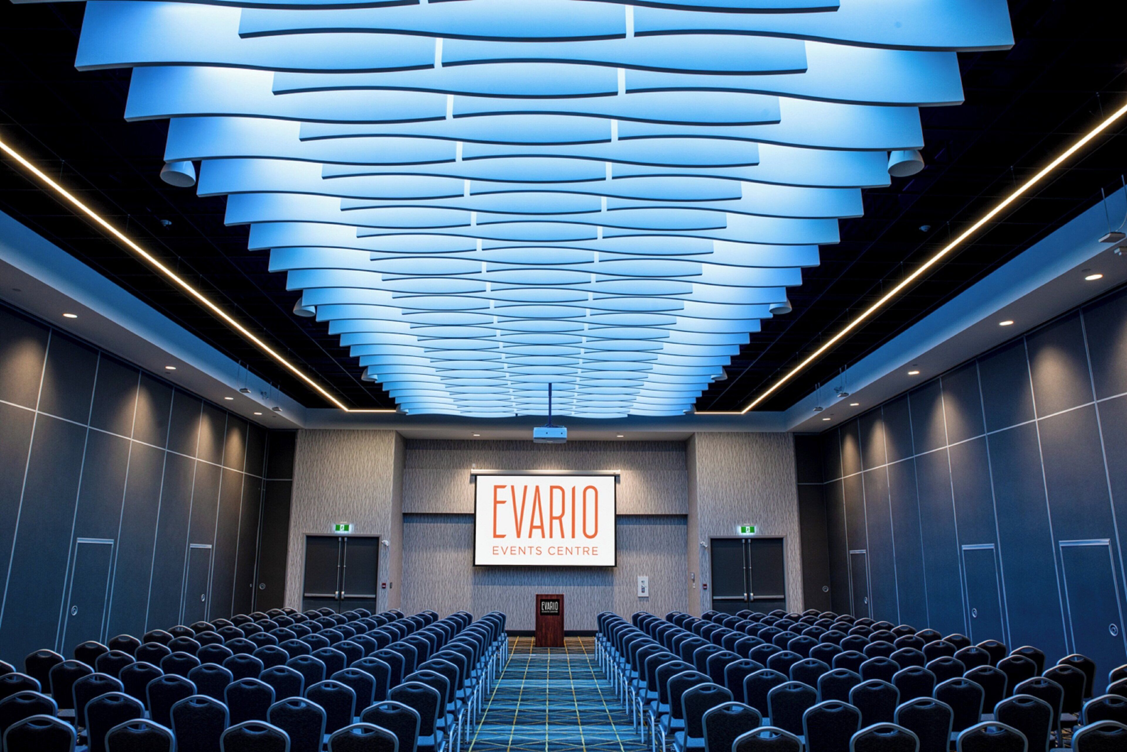 A conference room with rows of chairs, a large screen displaying 'EVARIO EVENTS CENTRE', and a modern ceiling design.
