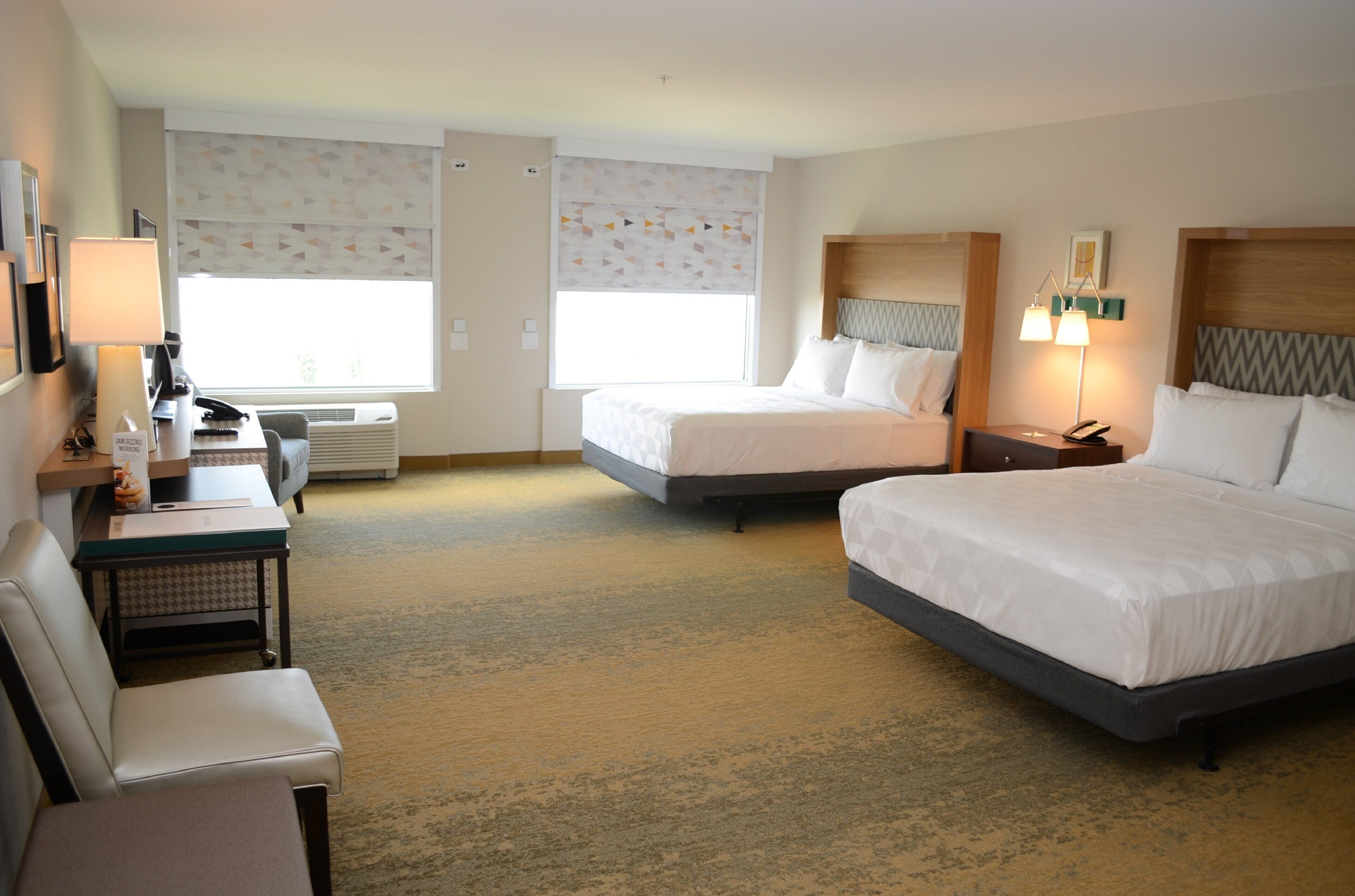 A hotel room with two beds, a desk with a chair, a bedside table with lamps, and a window with patterned curtains.