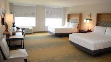 A hotel room with two beds, a desk with a chair, a bedside table with lamps, and a window with patterned curtains.
