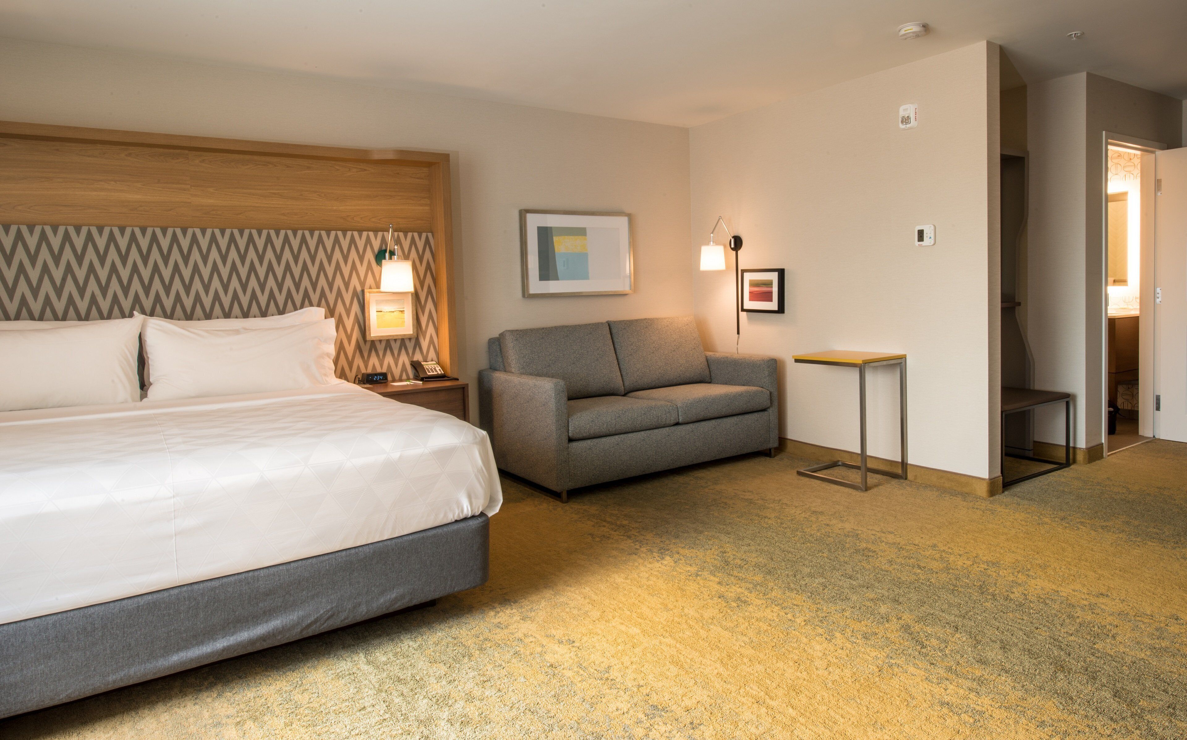 A hotel room with a large bed, a grey sofa, a small table, and a framed picture on the wall.
