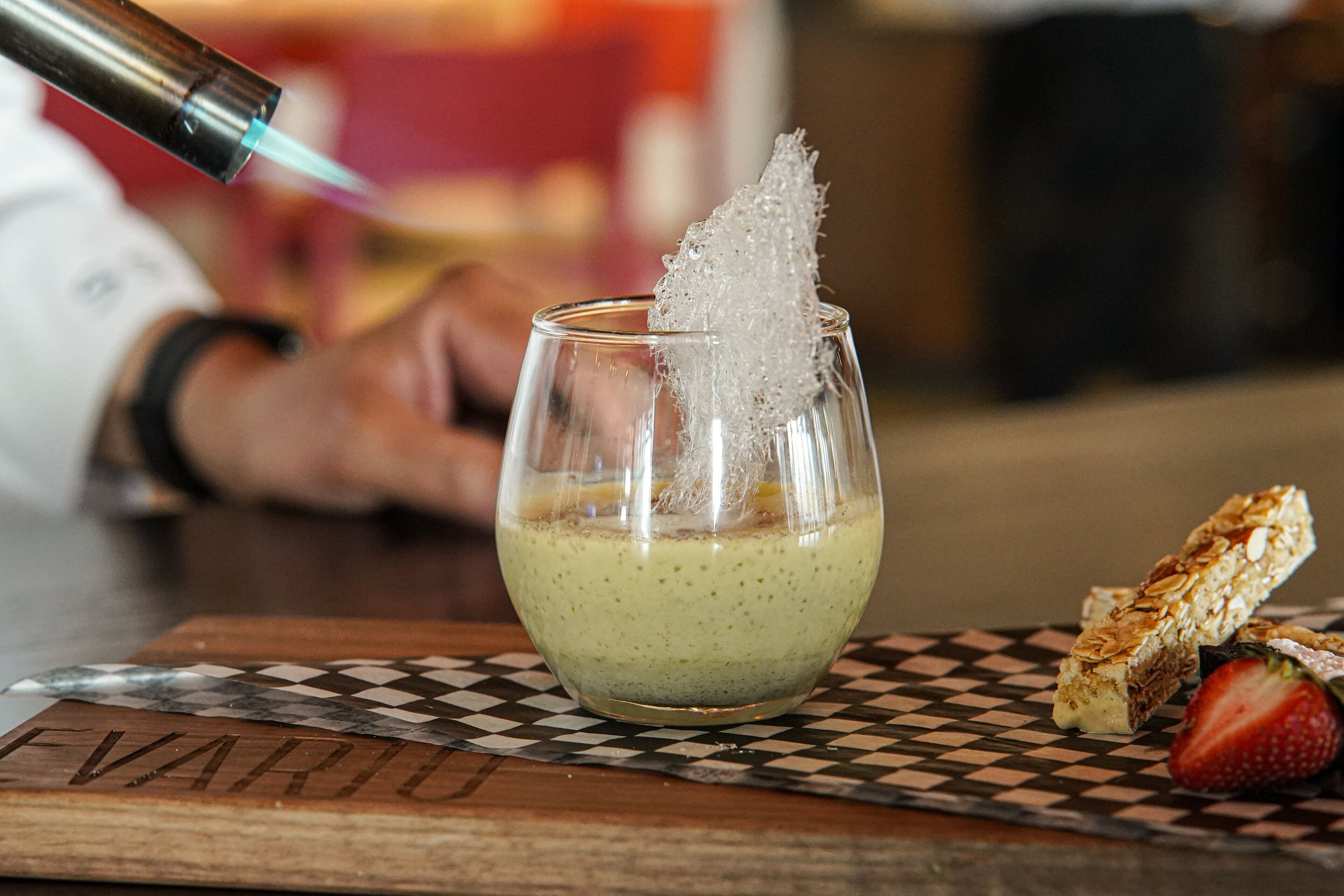 Een glas met een romig drankje, een suikerkristal en een stukje brood op een geruit kleed.