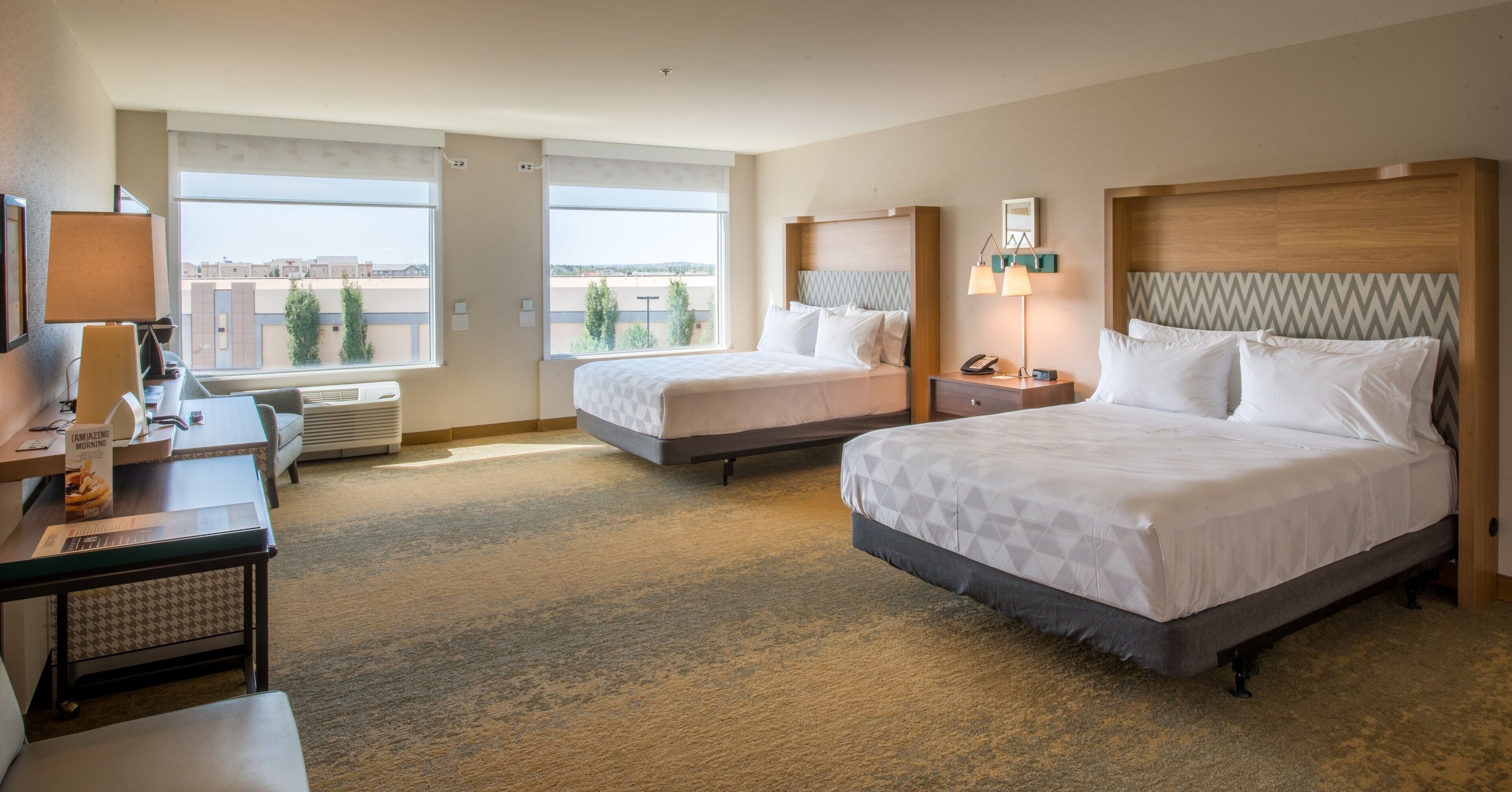 A hotel room with two beds, a desk, a chair, a lamp, and a view of the cityscape.