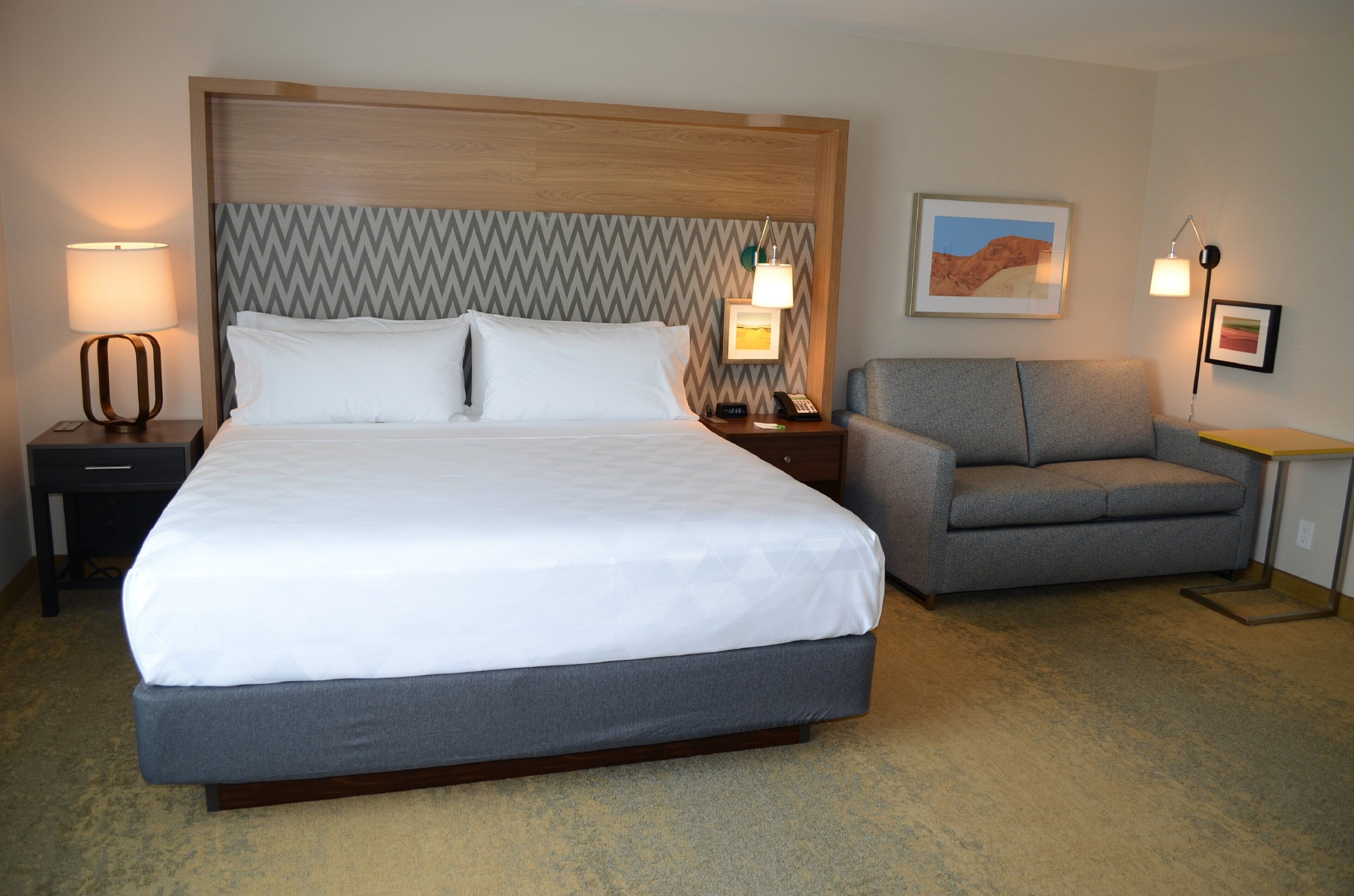 A hotel room with a large bed, a gray armchair, two bedside lamps, a nightstand, and a framed picture on the wall.