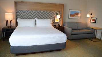 A hotel room with a large bed, a gray armchair, two bedside lamps, a nightstand, and a framed picture on the wall.