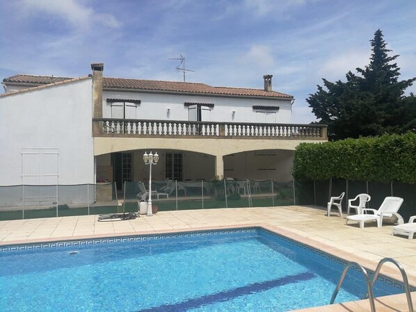 A swimming pool with a white building in the background, a covered patio area, and a few lounge chairs.