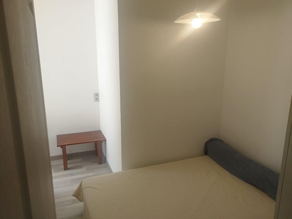 A small, sparsely furnished room with a bed, a bench, and a wall-mounted light fixture.