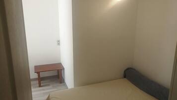 A small, sparsely furnished room with a bed, a bench, and a wall-mounted light fixture.
