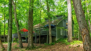 A cabin surrounded by tall trees, with a deck and a red umbrella. -  Coolfont 4 BDRM, lots of beds! Cozy Wooded Cottage. (Berkeley Springs)