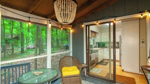 A cozy room with a large window overlooking a forest, a wicker chair with a yellow cushion, and a glass door leading to a kitchen. -  Coolfont 4 BDRM, lots of beds! Cozy Wooded Cottage. (Berkeley Springs)