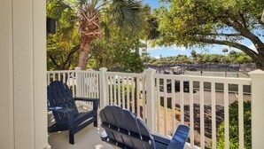 A covered outdoor area with a white fence, two Adirondack chairs, and a view of a tennis court and parking area. - Award-Winning Resort Amenities | Racquet Club Villa 23 (Isle of Palms)