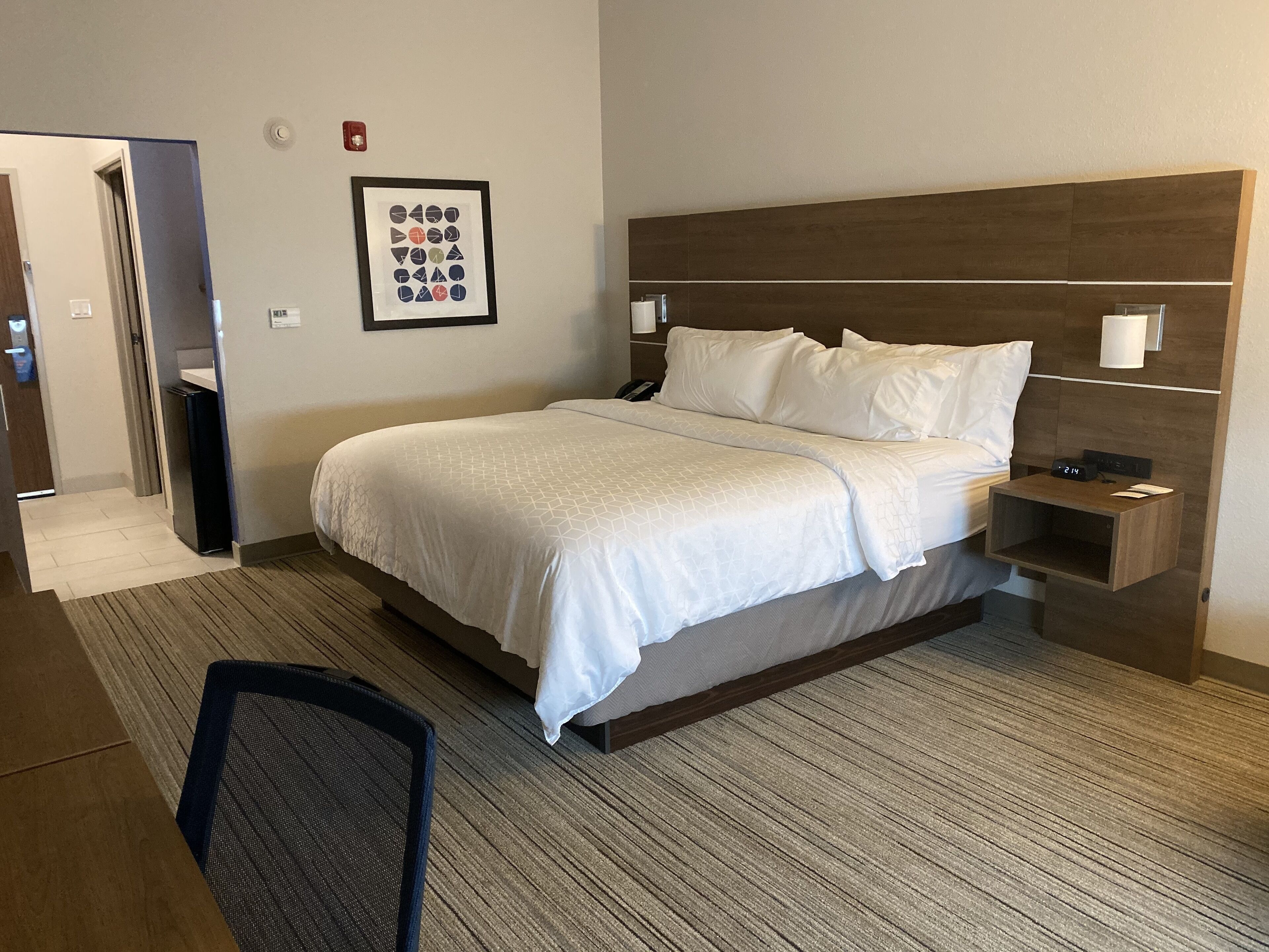 A hotel room with a bed, a nightstand, a chair, and a framed picture on the wall.