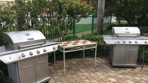 Two stainless steel barbecue grills with multiple burners, a metal table with a checkered tablecloth, and a paved patio area with a green fence and flowering bushes in the background. - Holiday Inn Express & Suites Junction by IHG (Junction)