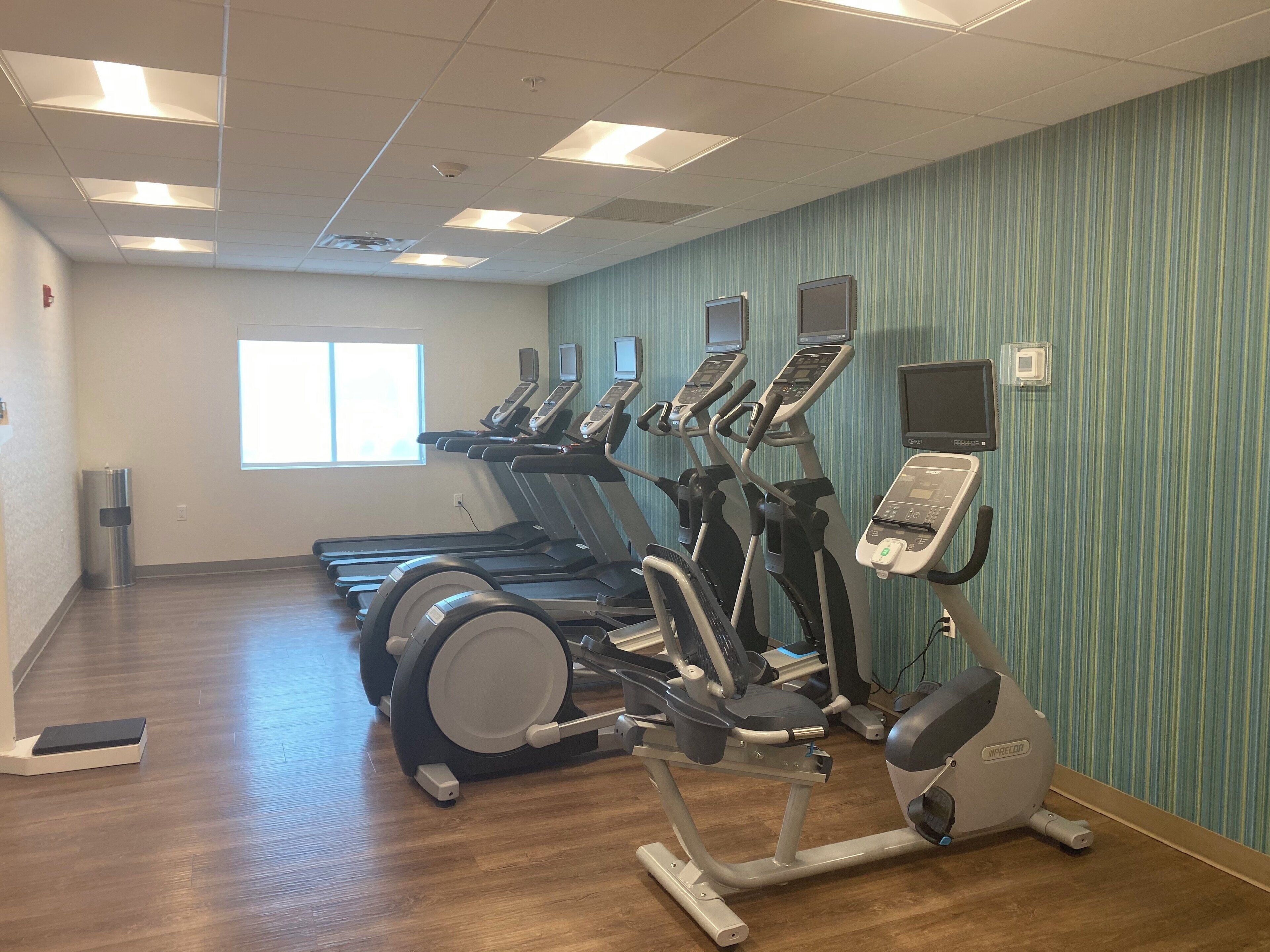 A fitness room with treadmills, elliptical machines, and a stationary bike, equipped with digital screens and a window providing natural light.