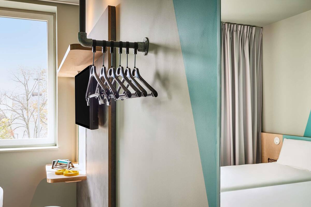 A hotel room with a bed, a window with a view of trees, a wall-mounted coat rack, and a small table with toiletries.