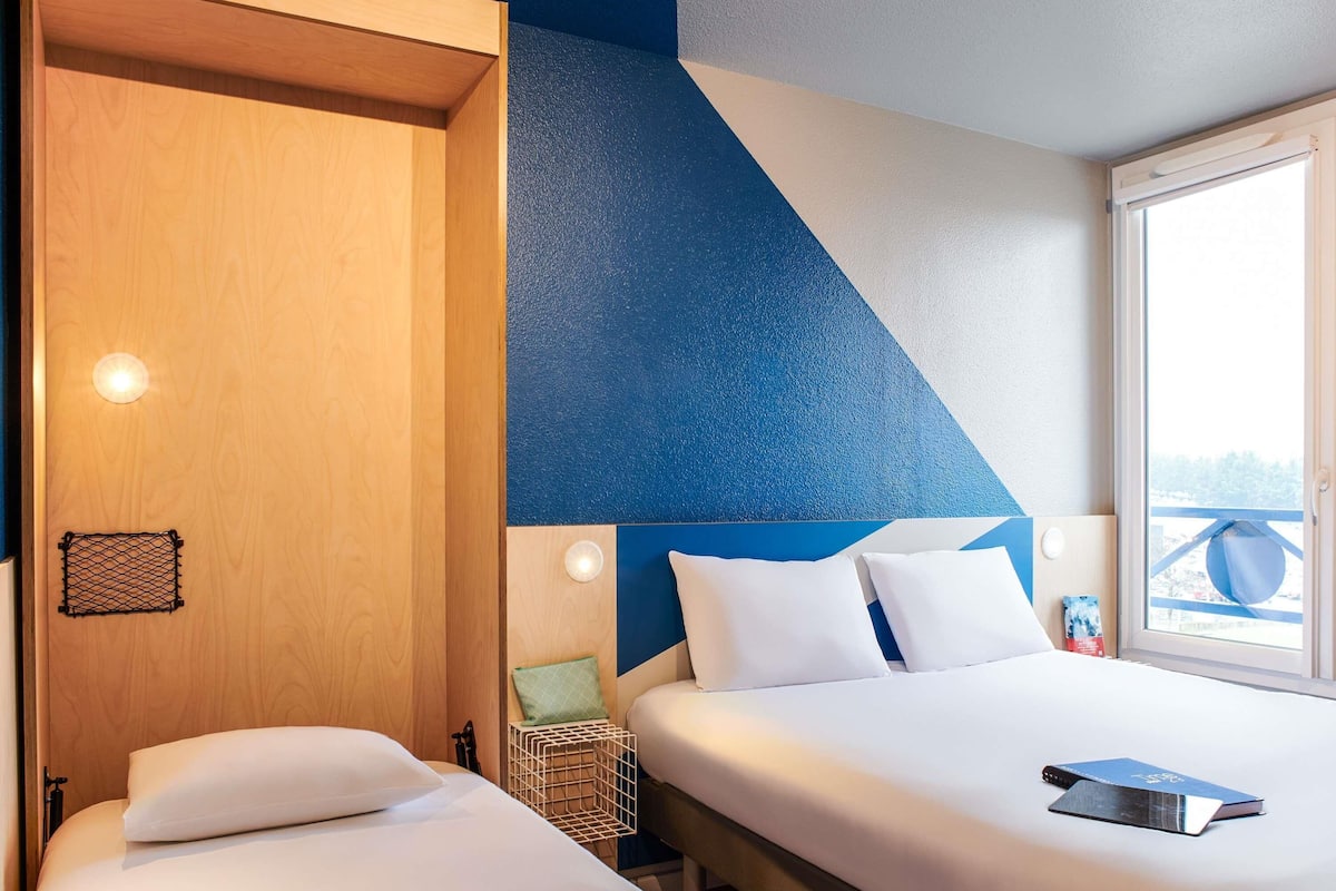 Une chambre d’hôtel avec deux lits, une porte en bois, un mur d’accent bleu et une fenêtre donnant sur un paysage agréable.