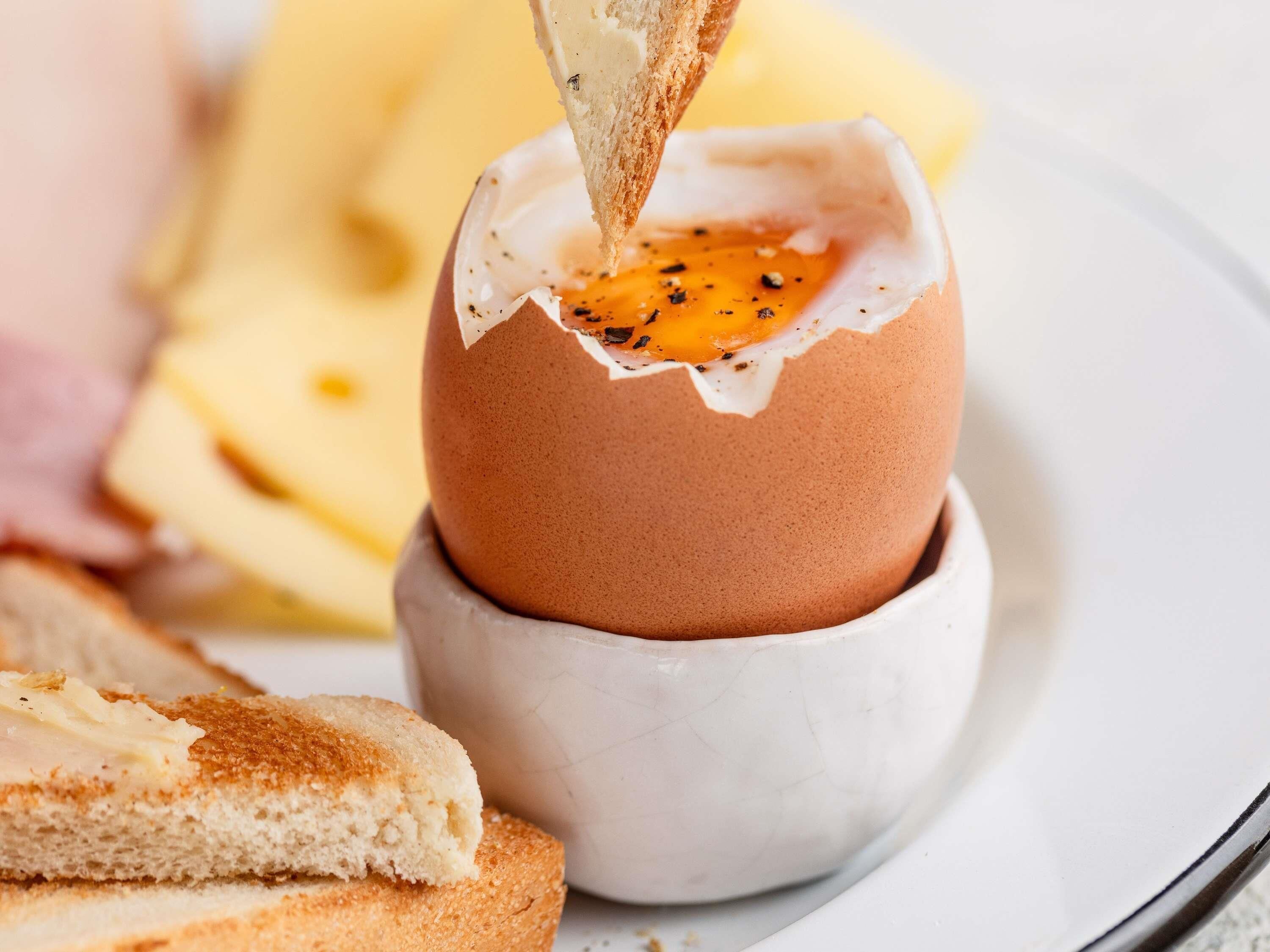 Een gebarsten eierdooier in een keramische kop, een sneetje toast met boter, en een bord met extra etenswaren.