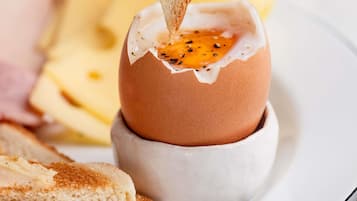 Een gebarsten eierdooier in een keramische kop, een sneetje toast met boter, en een bord met extra etenswaren.
