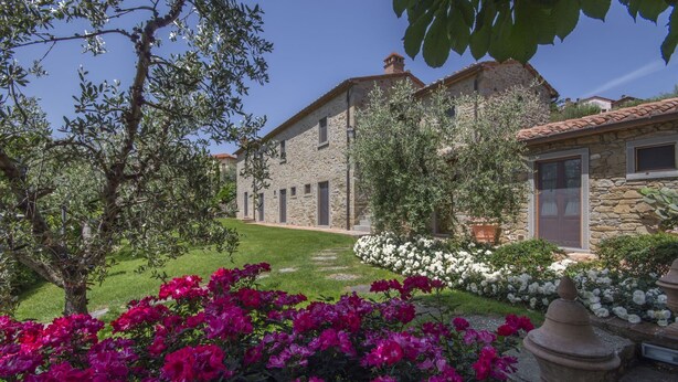 Un bâtiment en pierre doté d’un jardin où l’on trouve des fleurs roses et blanches, un sentier en pierre et un ciel d’un bleu limpide.