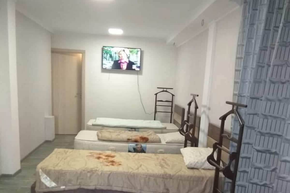 Ein Hotelzimmer mit drei Betten, einem an der Wand befestigten Fernseher und einem Fenster mit gemusterten Vorhängen.