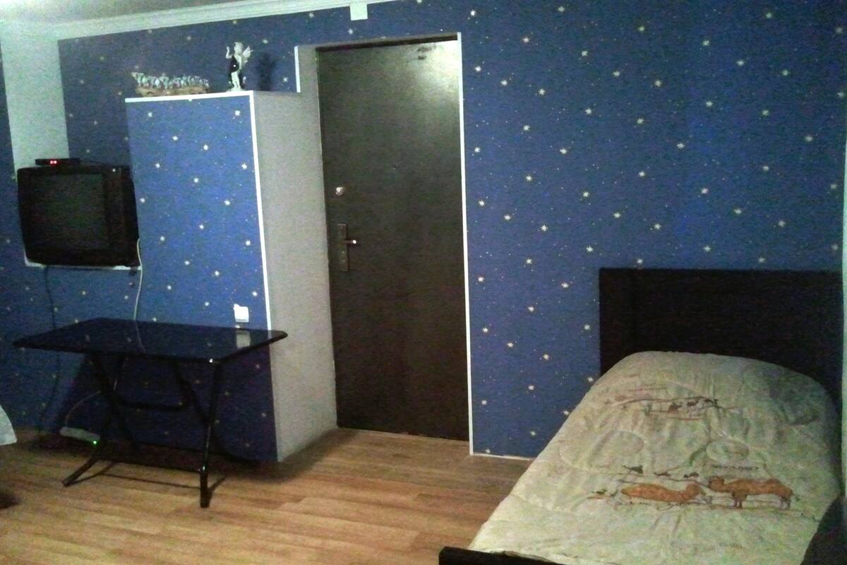 Ein Zimmer mit einer Wand im Sternenmuster, einem Bett mit Weltkarten-Design, einem Schreibtisch mit Computer und einer geschlossenen Tür.