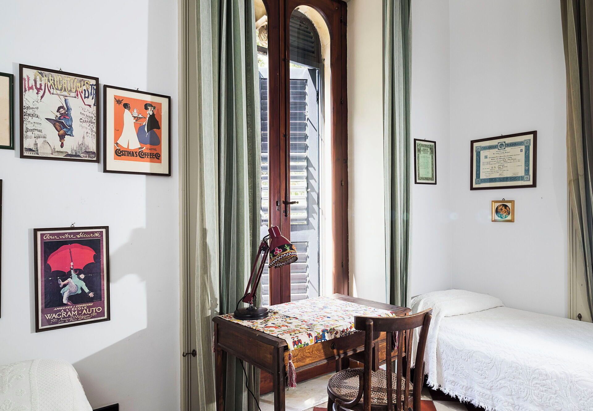 A room with a bed, a desk with a lamp, and various framed items on the walls.