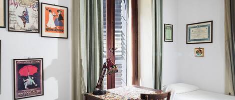 A room with a bed, a desk with a lamp, and various framed items on the walls.