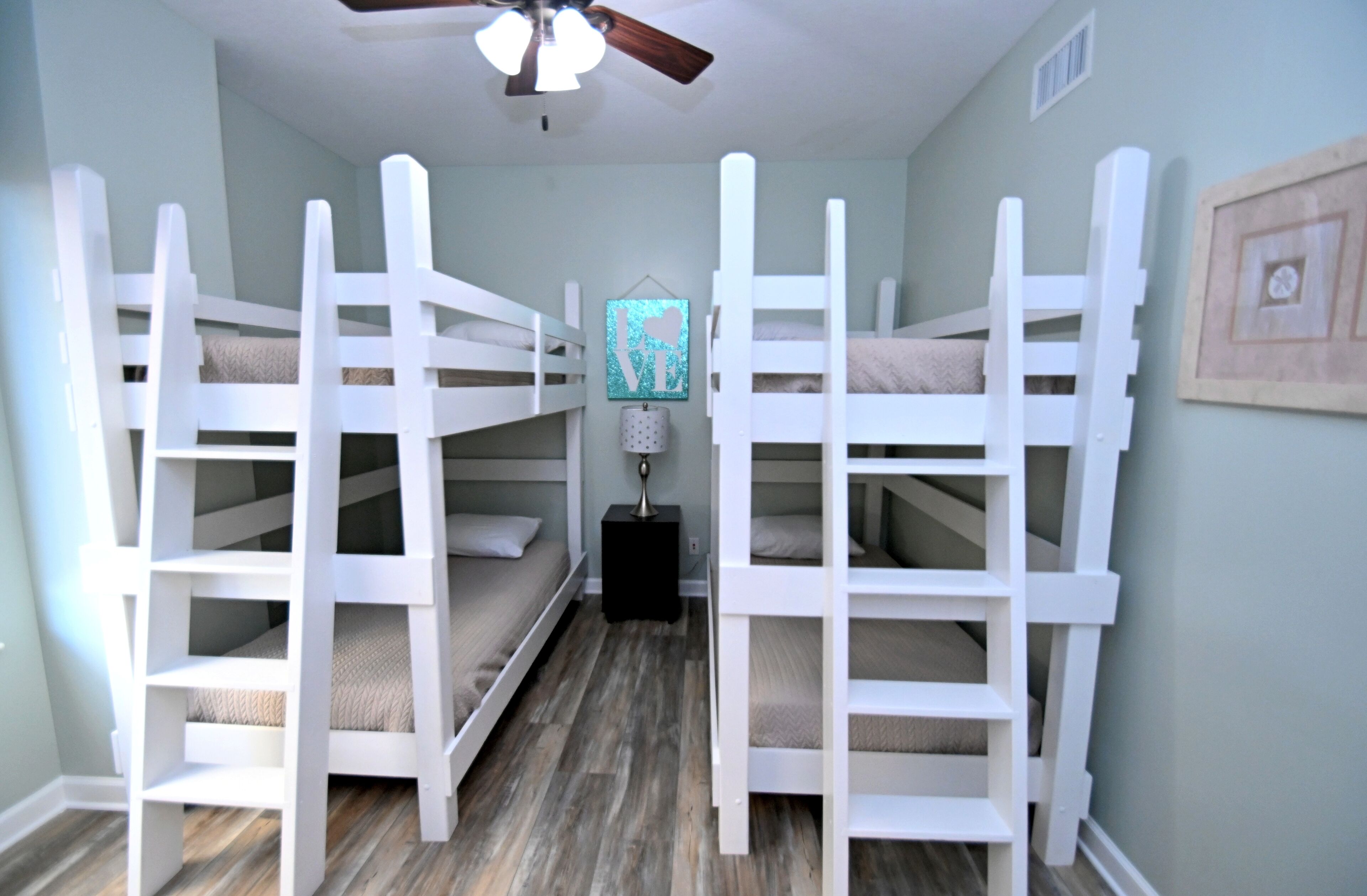 A room with bunk beds arranged in a U-shape, a ceiling fan, and a small table with a lamp.