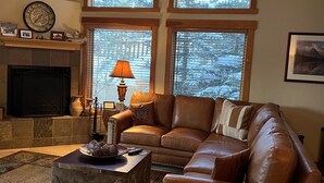 Una sala de estar acogedora con un sofá de cuero, una chimenea y grandes ventanales con vistas a árboles cubiertos de nieve.