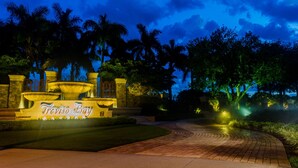 A well-lit entrance with a sign reading "Treviso Bay," surrounded by palm trees and a brick pathway.