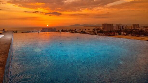 A swimming pool with a sunset view, surrounded by buildings and palm trees. - Casa Isacamil - Impeccable Views, Contemporary, Rooftop Pool, Proximity to Town (5 de Diciembre)