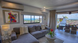 A modern living room with a grey sofa, a round coffee table with a vase of flowers, a city view through large windows, and a balcony with outdoor furniture. - Casa Isacamil - Impeccable Views, Contemporary, Rooftop Pool, Proximity to Town (5 de Diciembre)