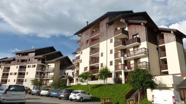 A multi-story hotel with balconies, a parking area, and a small hill with greenery.