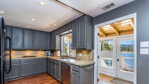 Una cocina moderna con armarios oscuros, encimeras de granito y lavavajillas. Cuenta con piso de madera y vista a un lago a través de una puerta de vidrio.