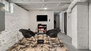 Una habitación con pared de ladrillo blanco, un televisor de pantalla plana sobre una estantería, y una alfombra estampada en el suelo.
