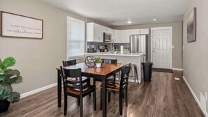 Una cocina moderna con armarios blancos, un refrigerador de acero inoxidable y un comedor con mesa y sillas de madera.