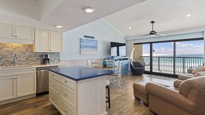 Een moderne keuken met witte kastjes, een eiland met blauw aanrecht en een vaatwasser. Er is een eethoek met een tafel en stoelen, en een balkon met uitzicht op de oceaan.