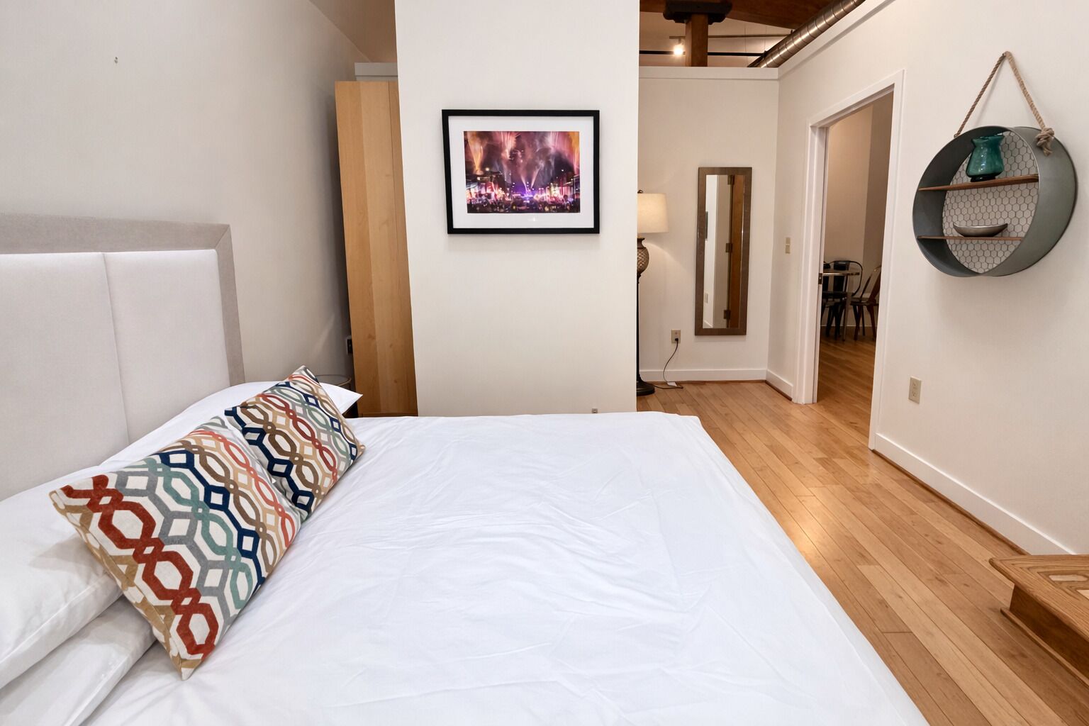Un dormitorio con una cama grande, un cojín colorido, un cuadro enmarcado en la pared y suelo de madera.