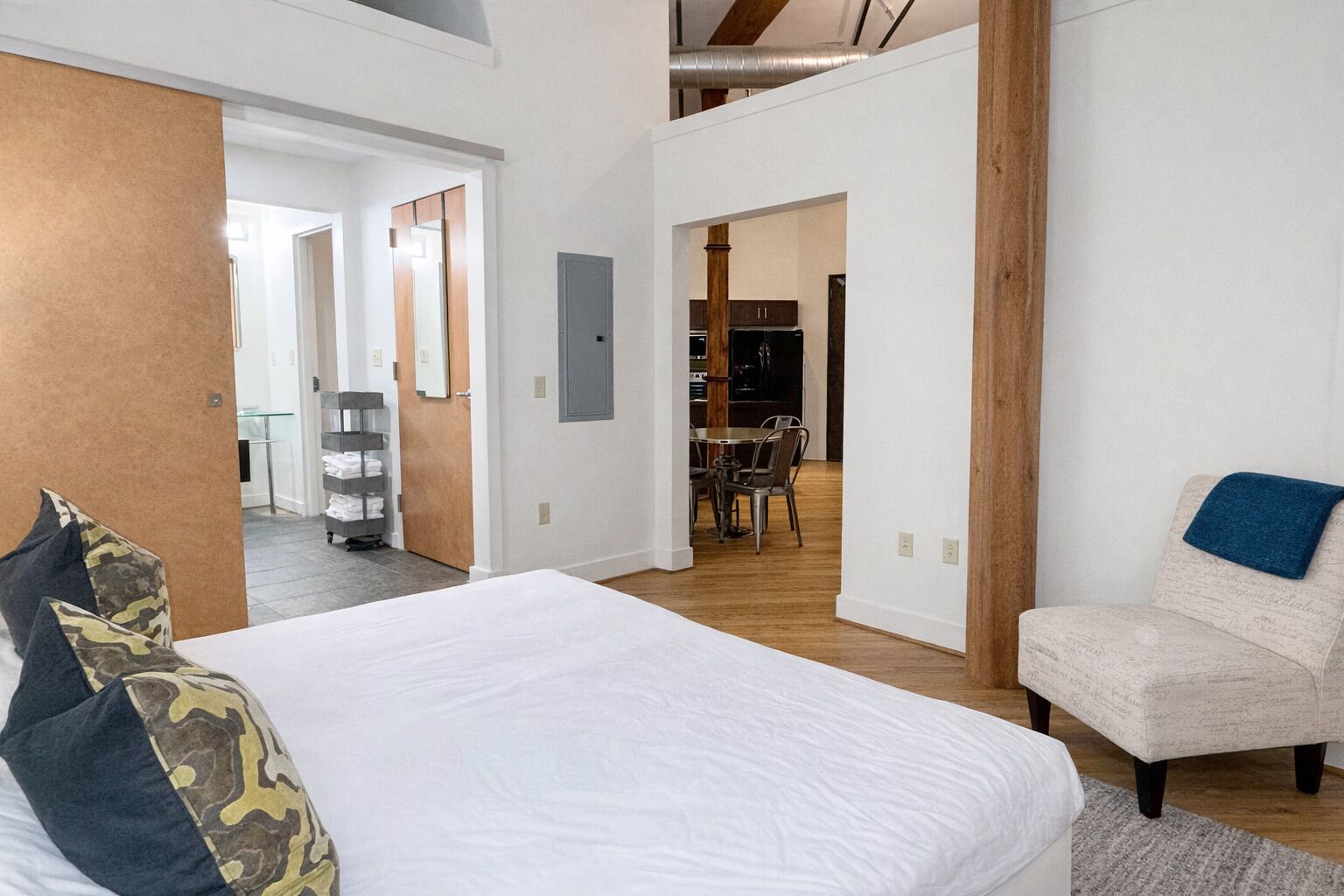 A bedroom with a large bed, a chair, and a view into the dining area.