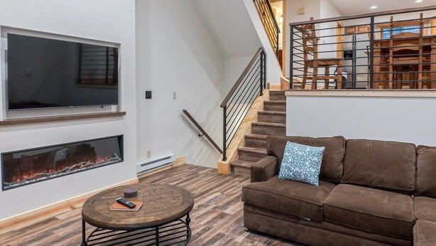 A modern living room with a large flat-screen TV, a fireplace, a brown sofa, and a staircase with a railing. - Walk to Skiing, Hiking and Main Street! (Breckenridge)