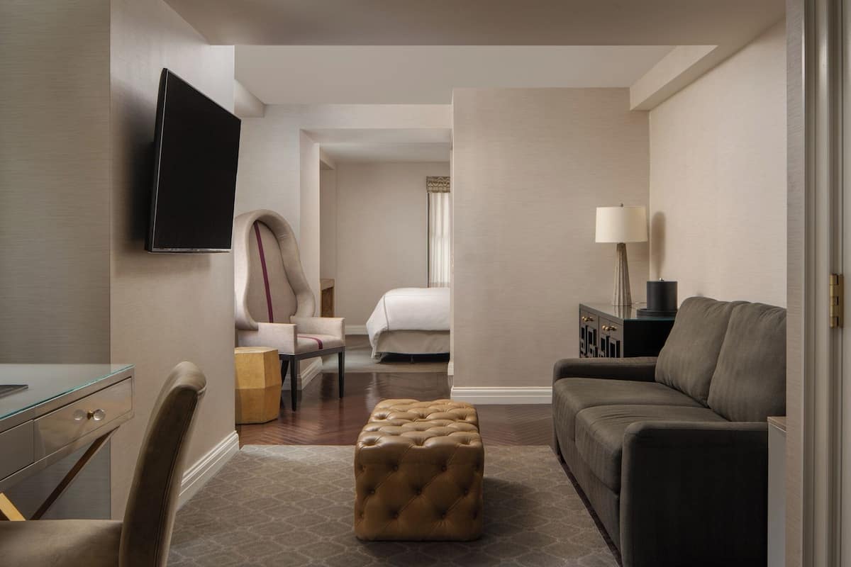 Une chambre d’hôtel équipée d’un téléviseur à écran plat fixé au mur, d’un pouf capitonné, d’un canapé, d’un bureau avec un plateau en verre, d’une chaise et d’une table de chevet avec une lampe.